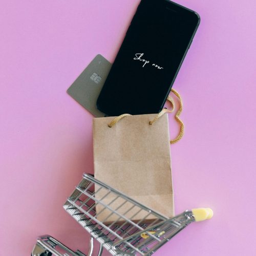 A smartphone, credit card, and bag in a miniature shopping cart on a pink background.